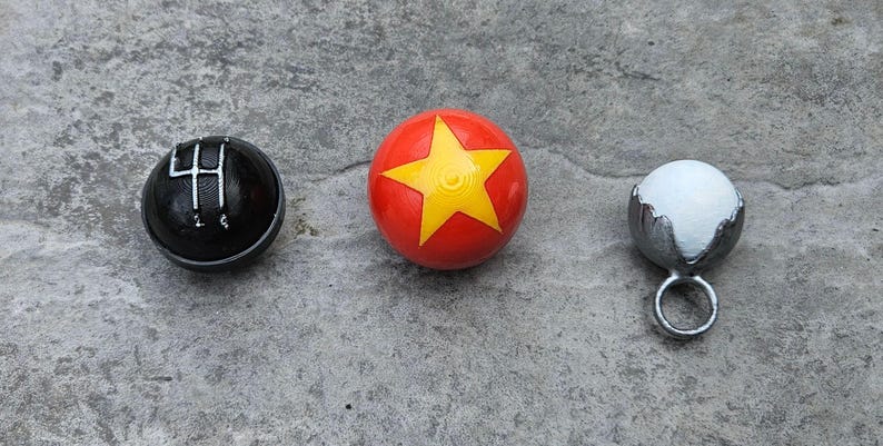 May include: Three small, round objects on a gray surface. A black shift knob with white markings, a red ball with a yellow star, and a silver-colored object with a white sphere inside.
