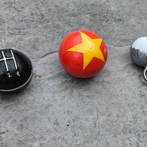 May include: Three spherical objects: a black shift knob with a silver gear pattern, a red ball with a yellow star, and a small white ball in a silver holder with a ring.