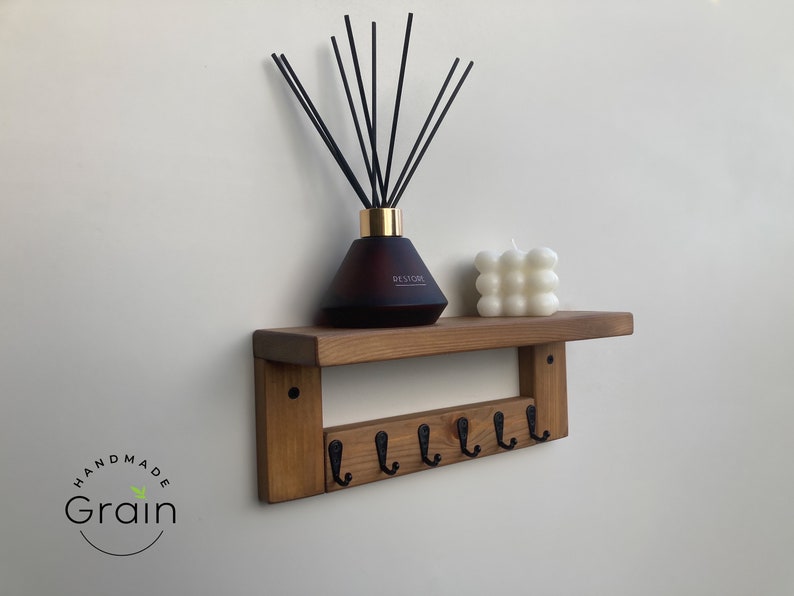 May include: A wooden shelf with six black hooks and a brown diffuser bottle with black sticks on top. A white candle is also on the shelf. The shelf is mounted on a white wall. The text "Handmade Grain" is visible in the lower left corner of the image.