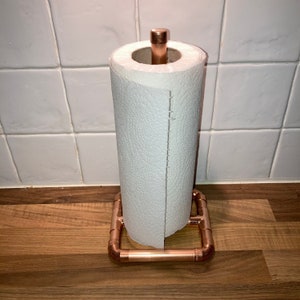 May include: A copper paper towel holder with a white paper towel roll. The holder has a square base and a round top.