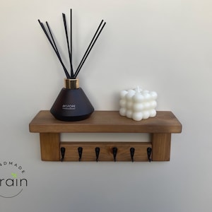May include: A wooden shelf with five hooks and a black diffuser bottle with black reeds and a white bubble candle on top. The shelf is mounted on a white wall. The text "Handmade Grain" is visible in the bottom left corner of the image.