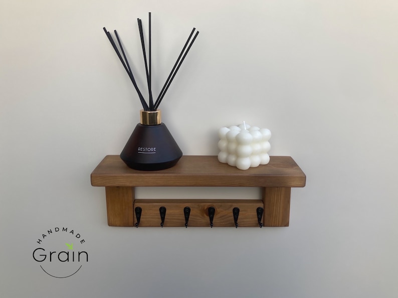 May include: A wooden shelf with five hooks and a black diffuser bottle with black reeds and a white bubble candle on top. The shelf is mounted on a white wall. The text "Handmade Grain" is visible in the bottom left corner of the image.
