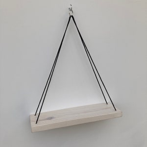 以下が含まれることがあります： 黒い紐で天井から吊り下げられた白い木製の棚。棚の長さは約30cm、幅は約10cmです。