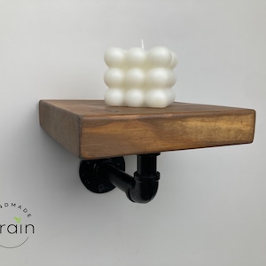 May include: A wooden shelf with black metal pipe brackets. The shelf is stained a dark brown color and has a white candle on top. The candle is shaped like a cube with rounded edges.