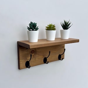 May include: A wall-mounted wooden shelf with a natural brown finish featuring three black metal hooks attached to the front panel. Three small artificial green succulent plants in white ceramic pots are arranged in a row on the top surface.