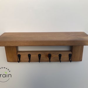 May include: A wooden shelf with a key rack underneath. The shelf is stained a dark brown color and has a rustic look. The key rack has six hooks and is made of wood. The shelf is labeled "Handmade Grain".