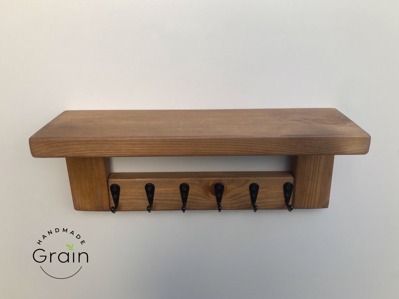 May include: A wooden shelf with a key rack underneath. The shelf is stained a dark brown color and has a rustic look. The key rack has six hooks and is made of wood. The shelf is labeled "Handmade Grain".