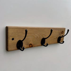 May include: A rectangular wooden wall rack featuring three matte black metal hooks. The wood has a natural light brown finish with visible grain patterns and knots. Each hook is secured with screws, providing a functional storage solution.