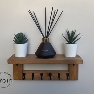 May include: A wooden shelf with five hooks and a brown diffuser bottle with black reeds. The shelf is decorated with two small potted succulents. The shelf is labeled "Handmade Grain".