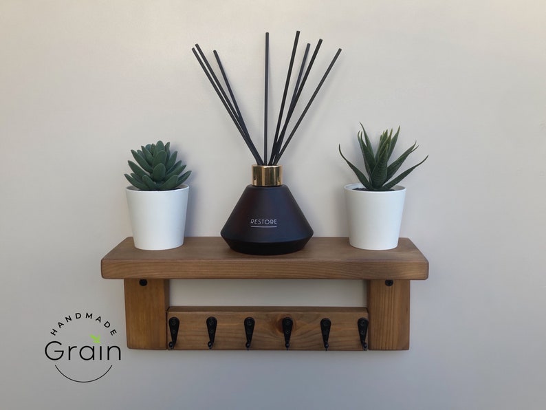 May include: A wooden shelf with five hooks and a brown diffuser bottle with black reeds. The shelf is decorated with two small potted succulents. The shelf is labeled "Handmade Grain".