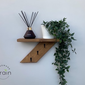 May include: A wooden shelf with a slanted design and three black hooks. The shelf is mounted on a white wall and has a dark brown finish. A black diffuser bottle with gold accents and a white planter with a green plant are on the shelf. The text "Handmade Grain" is visible in the lower left corner of the image.