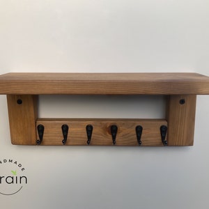 May include: A wooden key rack with a shelf on top. The rack has six black metal hooks and is stained a dark brown color. The shelf is made of the same wood and has a natural finish. The rack is labeled "Handmade Grain".