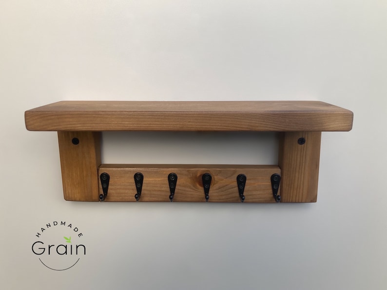 May include: A wooden key rack with a shelf on top. The rack has six black metal hooks and is stained a dark brown color. The shelf is made of the same wood and has a natural finish. The rack is labeled "Handmade Grain".