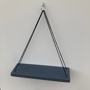 以下が含まれることがあります： 黒い紐で金属製のフックに吊るされた灰色の木製の棚。棚は長方形で、素朴な仕上げになっています。