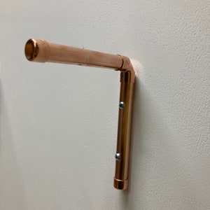 May include: A copper pipe shelf bracket with a 90-degree angle. The bracket is mounted to a white wall with two screws.
