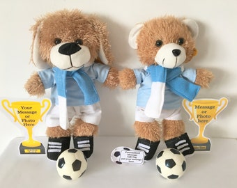 May include: Two plush toys, a dog and a bear, wearing blue and white soccer uniforms. Both toys are holding a soccer ball and standing next to a trophy with a blank space for a message or photo. The toys are on a white surface.