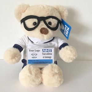 May include: A cream-coloured plush teddy bear wearing black plastic glasses and a white shirt with navy blue trim. The bear has a clear plastic ID badge attached to its chest that reads NHS, Your Logo, Dr Ted, and Looking After My Patients.