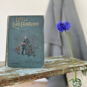 May include: A vintage hardcover book titled "Little Lord Fauntleroy" by Frances Hodgson Burnett. The book cover features a blue background with gold lettering and an illustration of a boy, a man, and a dog in a room.