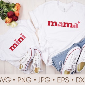May include: Two white t-shirts with red and pink text. One shirt says "mama" with a small heart and the other says "mini" with two small hearts. The shirts are laid out on a white wooden surface with a pair of white sneakers and blue jeans.