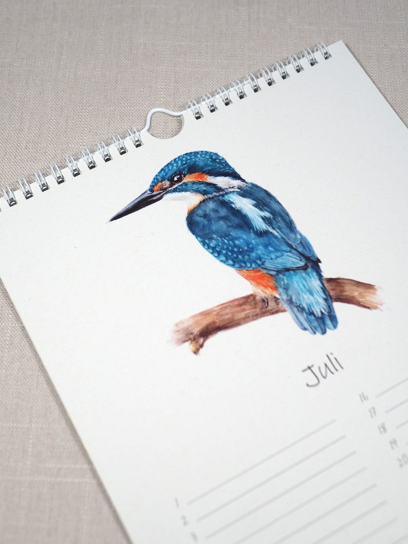 May include: A watercolour illustration of a blue and orange kingfisher perched on a branch. The illustration is on a white calendar page with the word "Juli" written in black ink. The calendar page has a white spiral binding.