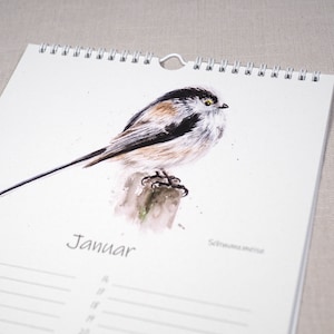 May include: A watercolour illustration of a long-tailed tit bird perched on a branch. The bird is brown, black, and white with a yellow eye. The illustration is on a white calendar page with the word "Januar" printed in black.