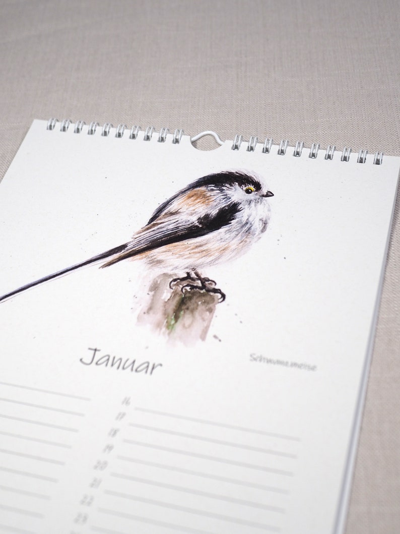 May include: A watercolour illustration of a long-tailed tit bird perched on a branch. The bird is brown, black, and white with a yellow eye. The illustration is on a white calendar page with the word "Januar" printed in black.