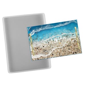 May include: A rectangular resin tray with a beach scene design. The tray features a vibrant blue ocean, white foamy waves, and a sandy shore with small pebbles. It has gold-coloured handles and a clear, rectangular mould.