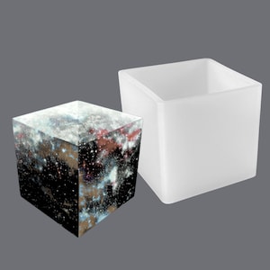 May include: Two square silicone moulds, one filled with a galaxy-themed resin cube and the other empty. The moulds are white and have a smooth, matte finish.