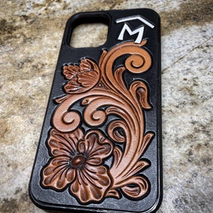 Peut inclure: Étui de téléphone en cuir noir avec un motif floral brun ciselé et la lettre "M" en blanc sur le dessus.