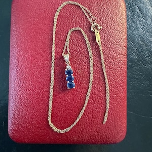 May include: A gold chain necklace featuring a vertical pendant with three oval-cut blue sapphire stones arranged in a row, topped with a single small clear accent stone. The necklace includes a spring ring clasp and a small rectangular metal tag.