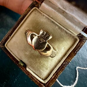 May include: A gold Claddagh ring, with a heart, crown, and hands design, is displayed in a vintage jewellery box with a velvet interior. The ring is a symbol of love, loyalty, and friendship.