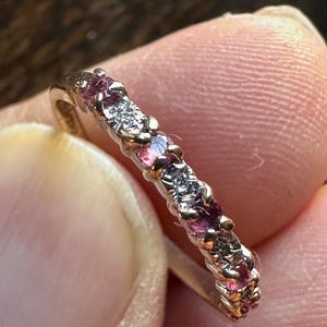 May include: A silver ring featuring alternating pink and clear gemstones. The ring is held between fingers, highlighting the intricate setting and the sparkle of the stones. The gemstones are small and evenly spaced around the band.