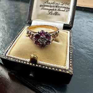 Op de afbeelding: Een vintage gouden ring met een cluster van rode edelstenen in een bloemmotief. De ring wordt gepresenteerd in een zwarte en gouden sieradendoos met de tekst "Prestons Ltd" aan de binnenkant. Het ontwerp is gedetailleerd.