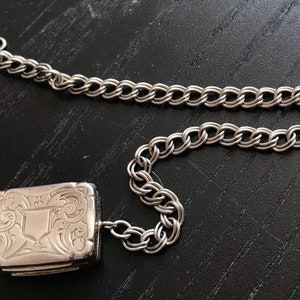 Op de afbeelding: Een zilveren medaillon met een sierlijk ontwerp en een zilveren ketting. Het medaillon is rechthoekig en heeft een scharnierend deksel.
