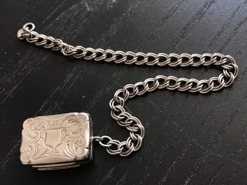 Op de afbeelding: Een zilveren medaillon met een sierlijk ontwerp en een zilveren ketting. Het medaillon is rechthoekig en heeft een scharnierend deksel.