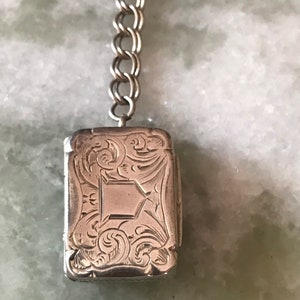 Op de afbeelding: Een zilveren medaillon met een sierlijk gegraveerd ontwerp hangt aan een zilveren ketting. Het medaillon is rechthoekig en heeft een scharnierend deksel.
