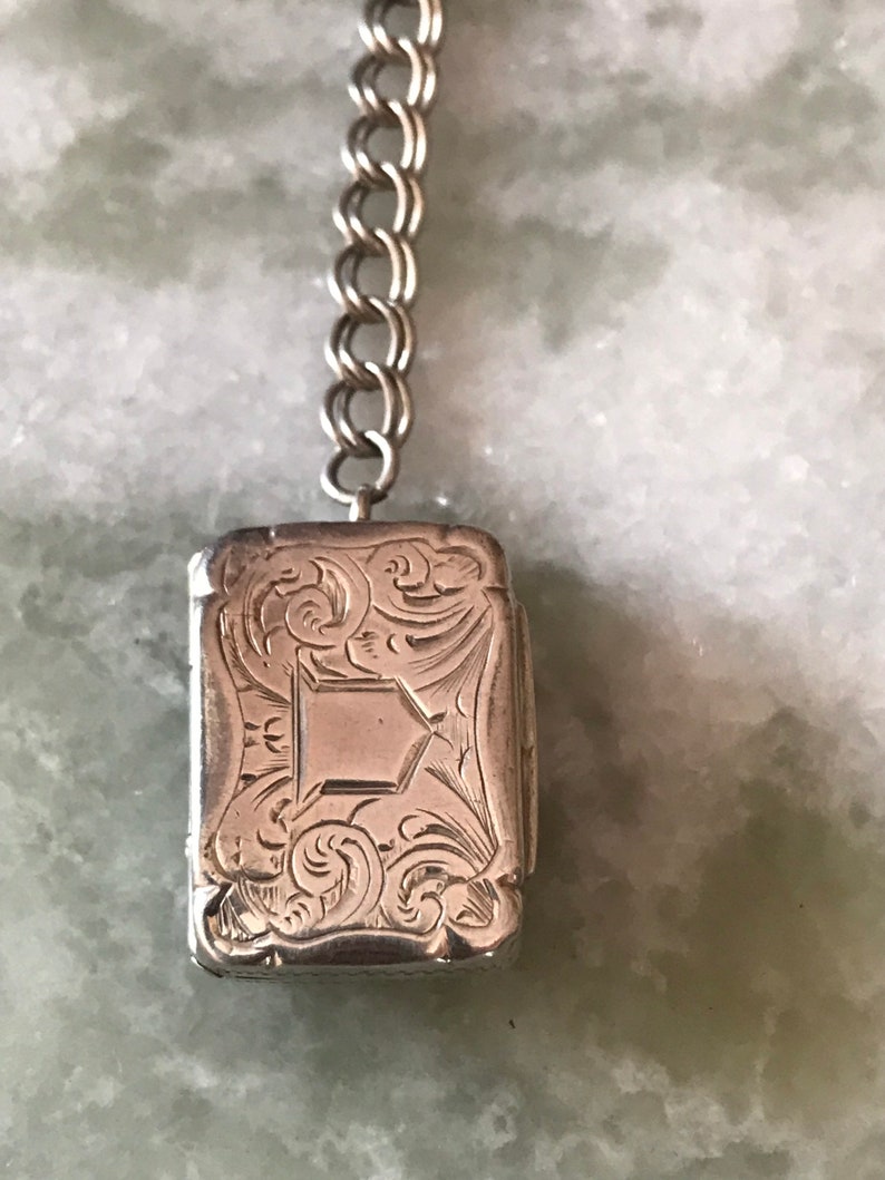 Op de afbeelding: Een zilveren medaillon met een sierlijk gegraveerd ontwerp hangt aan een zilveren ketting. Het medaillon is rechthoekig en heeft een scharnierend deksel.
