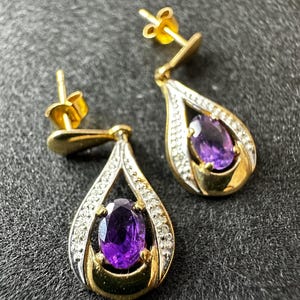 Puede incluir: Un par de pendientes dorados en forma de lágrima con piedras preciosas ovaladas de color púrpura intenso. Los pendientes tienen un halo de pequeñas piedras transparentes alrededor de las gemas púrpuras.