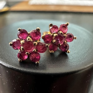 Könnte beinhalten: Ein Paar Ohrstecker in Blumenform mit sechs runden, tiefrosa facettierten Edelsteinen, die einen zentralen Stein umgeben, eingefasst in einen goldfarbenen Metallrahmen, wobei jeder einzelne Stein sicher von Krappenfassungen gehalten wird.