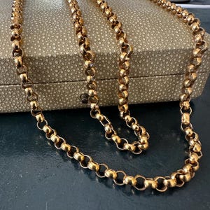 以下が含まれることがあります： 金色のチェーンネックレス、ロロチェーンのデザイン。 ネックレスは、質感のある明るい色の箱の上に掛けられています。 チェーンは、連動する円形のリンクでできており、クラシックでエレガントな外観を作り出しています。