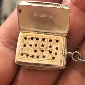 Op de afbeelding: Een zilveren medaillon met een scharnierend deksel en een decoratief, geperforeerd ontwerp. Het medaillon heeft een ketting en de binnenkant van het deksel is gemarkeerd met de letters "T & J".