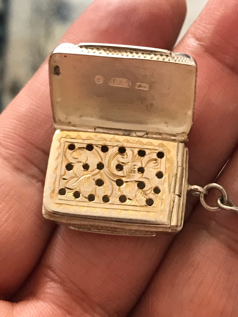 Op de afbeelding: Een zilveren medaillon met een scharnierend deksel en een decoratief, geperforeerd ontwerp. Het medaillon heeft een ketting en de binnenkant van het deksel is gemarkeerd met de letters "T & J".