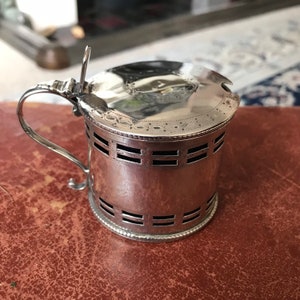 May include: A silver sugar or creamer jar with a hinged lid and a decorative handle. The jar has a patterned design around the top and bottom and a series of horizontal lines around the middle.