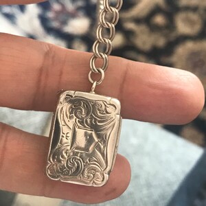 Op de afbeelding: Een zilveren medaillon met een ingewikkeld bloemmotief en een diamantvormig midden. Het medaillon is bevestigd aan een zilveren ketting.