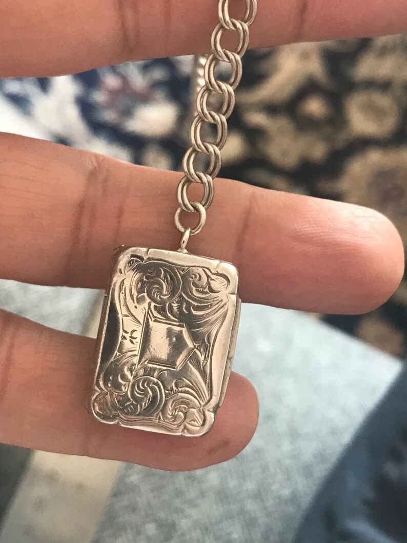 Op de afbeelding: Een zilveren medaillon met een ingewikkeld bloemmotief en een diamantvormig midden. Het medaillon is bevestigd aan een zilveren ketting.