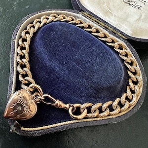 Puede incluir: Un brazalete dorado con un relicario en forma de corazón, en una caja de joyería de terciopelo azul. El brazalete presenta un diseño de cadena gruesa. El relicario tiene un pequeño diamante y grabados decorativos. La caja está abierta.