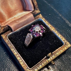 May include: A vintage ring with a floral design, featuring a central diamond surrounded by vibrant purple gemstones. The ring is set in a silver band with a textured design. It rests inside a velvet-lined, antique jewelry box with a decorative gold trim.