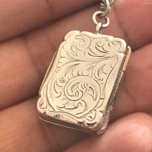 Op de afbeelding: Een zilveren medaillon met een ingewikkeld bloemmotief. Het medaillon is gesloten en heeft een ketting eraan.