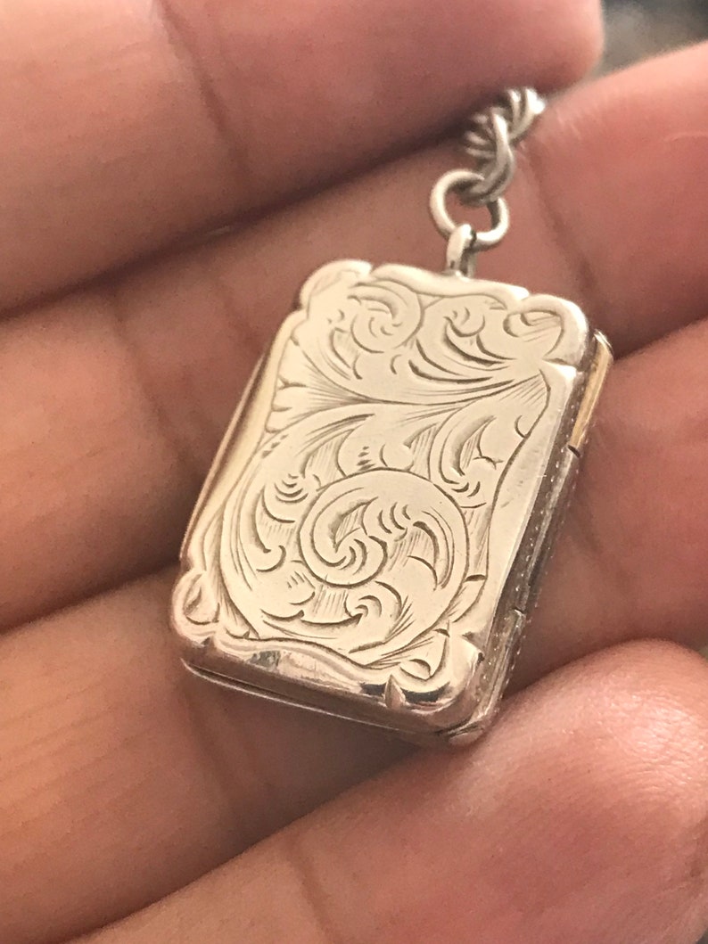 Op de afbeelding: Een zilveren medaillon met een ingewikkeld bloemmotief. Het medaillon is gesloten en heeft een ketting eraan.