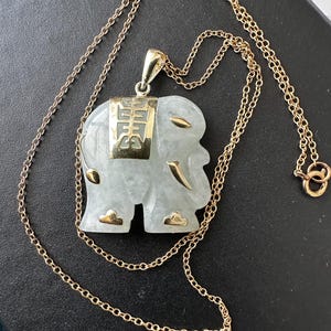 Puede incluir: Collar de cadena dorada con un colgante de elefante. El colgante es de color verde claro con detalles dorados y una placa rectangular dorada con caracteres chinos. La cadena tiene un diseño delicado.